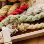 Yarn Tools - From above of unfinished tapestry on manual wooden loom placed on wooden table with assorted yarn twists and wooden needles during weaving process in workshop