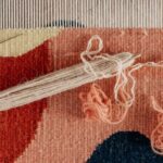 Yarn Tools - From above of shuttle and needle placed on part of handmade carpet with circle pattern on loom frame during weaving process